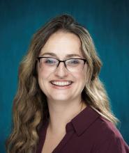 Brunette with long hair and glasses, wearing a purple shirt smiles at the camera