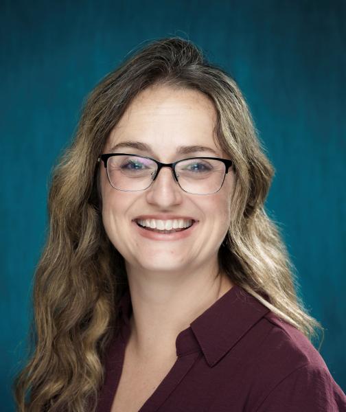 Brunette with long hair and glasses, wearing a purple shirt smiles at the camera